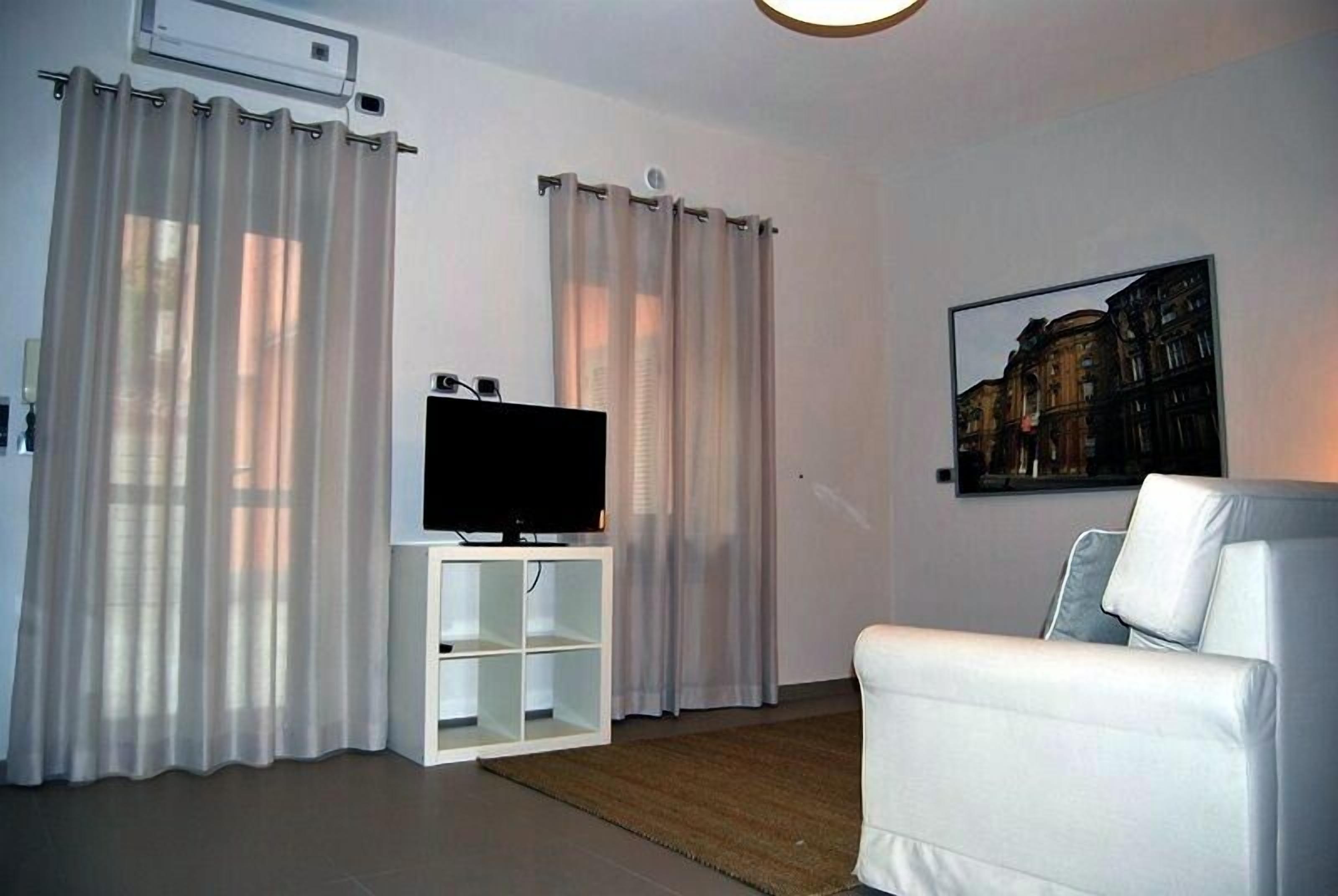 Un salon moderne avec un téléviseur à écran plat posé sur un meuble blanc, des rideaux blancs, un canapé blanc et un tableau représentant un paysage urbain, encadré et accroché au mur.