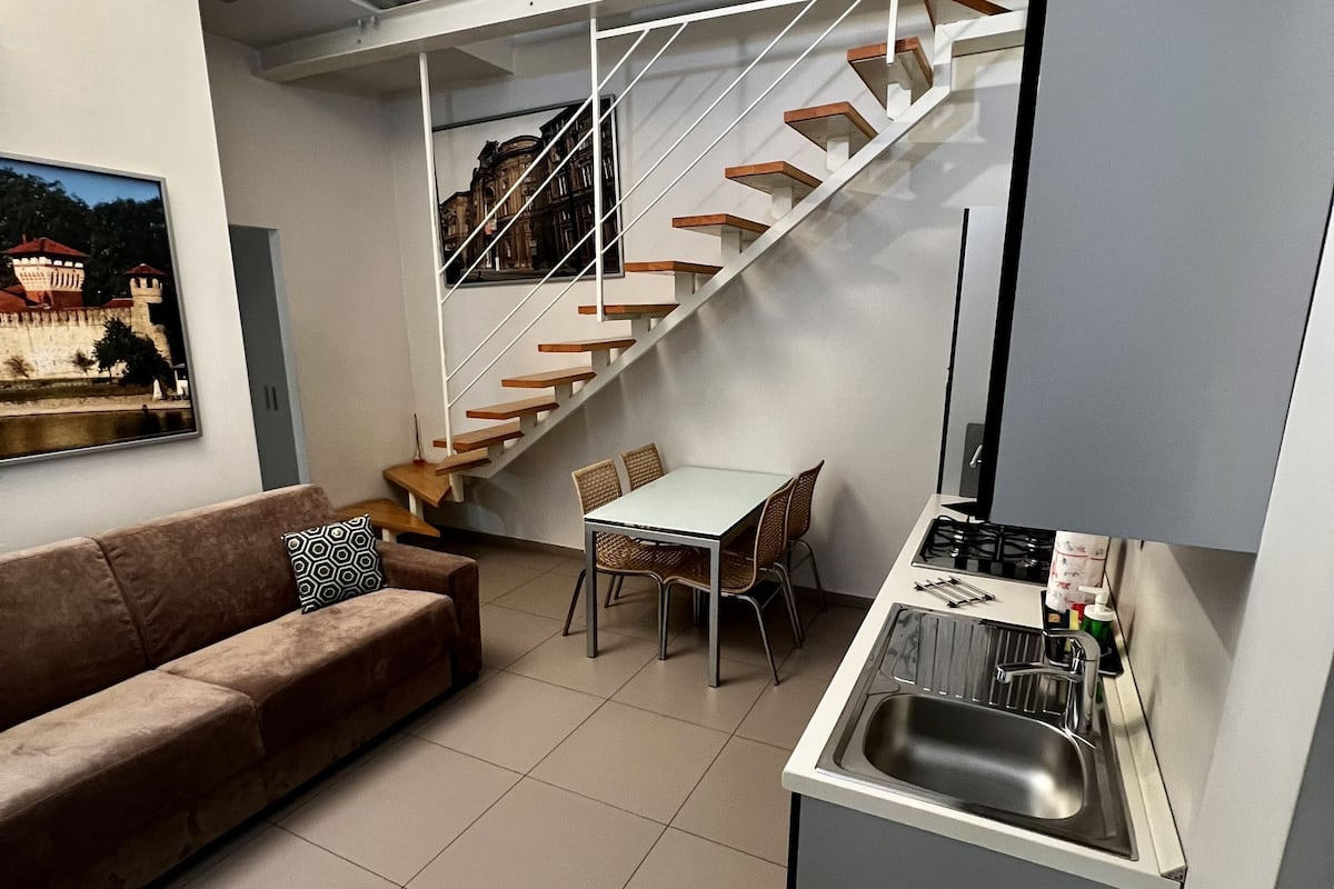 Een compacte keuken met een ingebouwde kookplaat, een spoelbak en een eethoek met een tafel en stoelen. Een trap leidt naar een hoger gelegen verdieping.