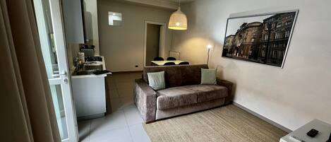 A modern apartment with a kitchenette, a sofa, a dining area, and a large window with a cityscape view.