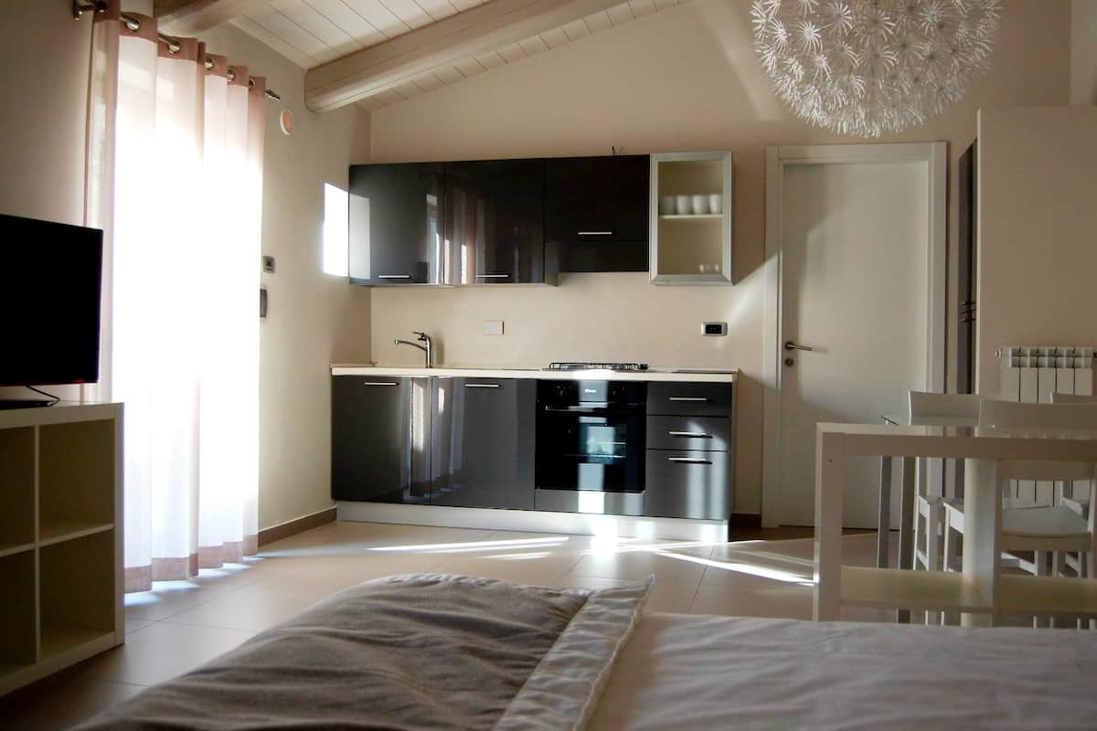 Een moderne keuken met roestvrijstalen apparatuur, een ingebouwde oven en een magnetron. Links staat een tv op een standaard, en op de voorgrond staat een witte stoel.