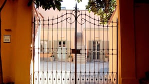 Un portail en fer forgé, agrémenté d’un motif décoratif sur le dessus, mène à un bâtiment dont la façade est de couleur claire et les fenêtres sont dotées de volets.
