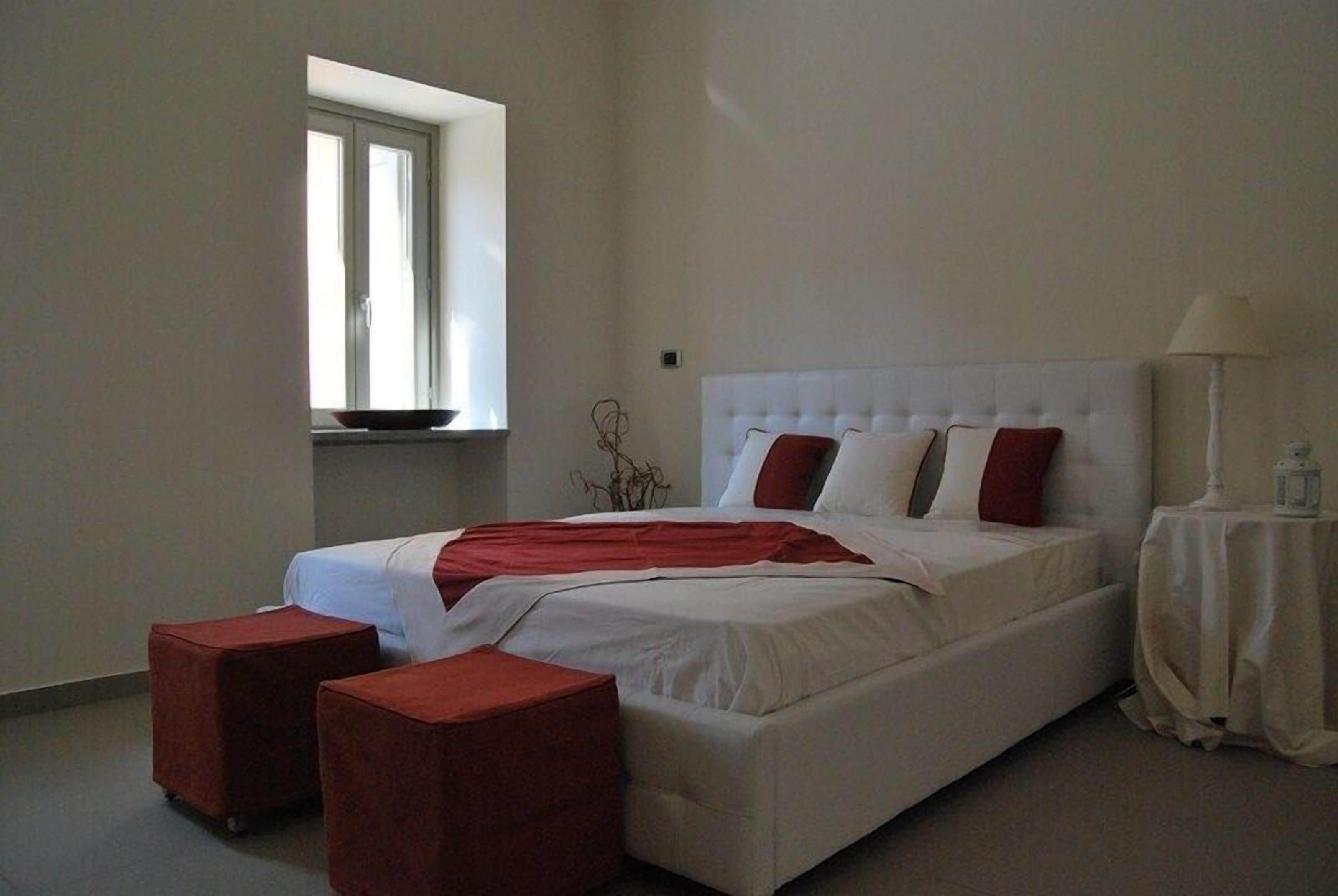 Une chambre à coucher avec un grand lit, deux poufs rouges, une fenêtre et une table de chevet avec une lampe.