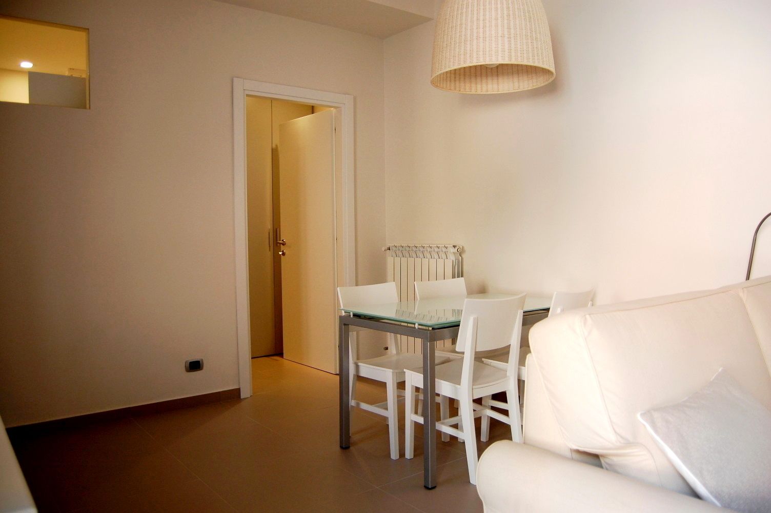 A modern living room with a white sofa, a glass dining table, and a white chair. There is a door leading to another room and a radiator on the wall.