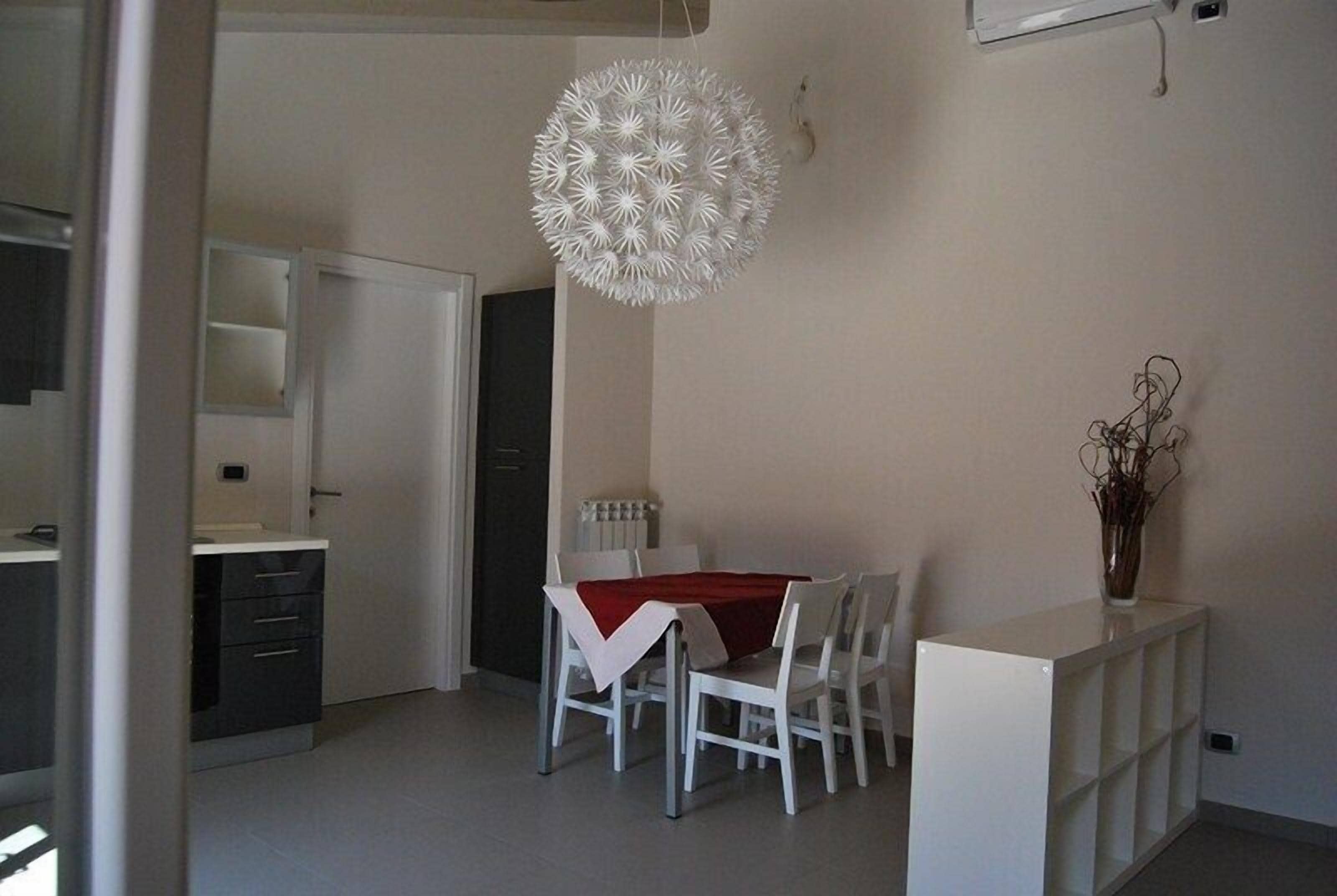 A dining area with a white table, red tablecloth, and white chairs. There is a white cabinet with a vase on top.
