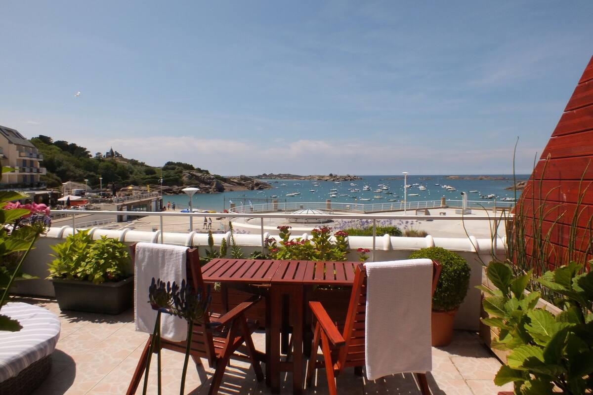 A wooden table and chairs on a terrace with a view of a coastal area, including boats and a rocky shoreline.