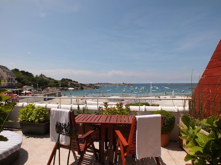 Une table et des chaises en bois sur une terrasse offrant une vue sur une zone côtière, avec des bateaux et une côte rocheuse.. Le Beauséjour