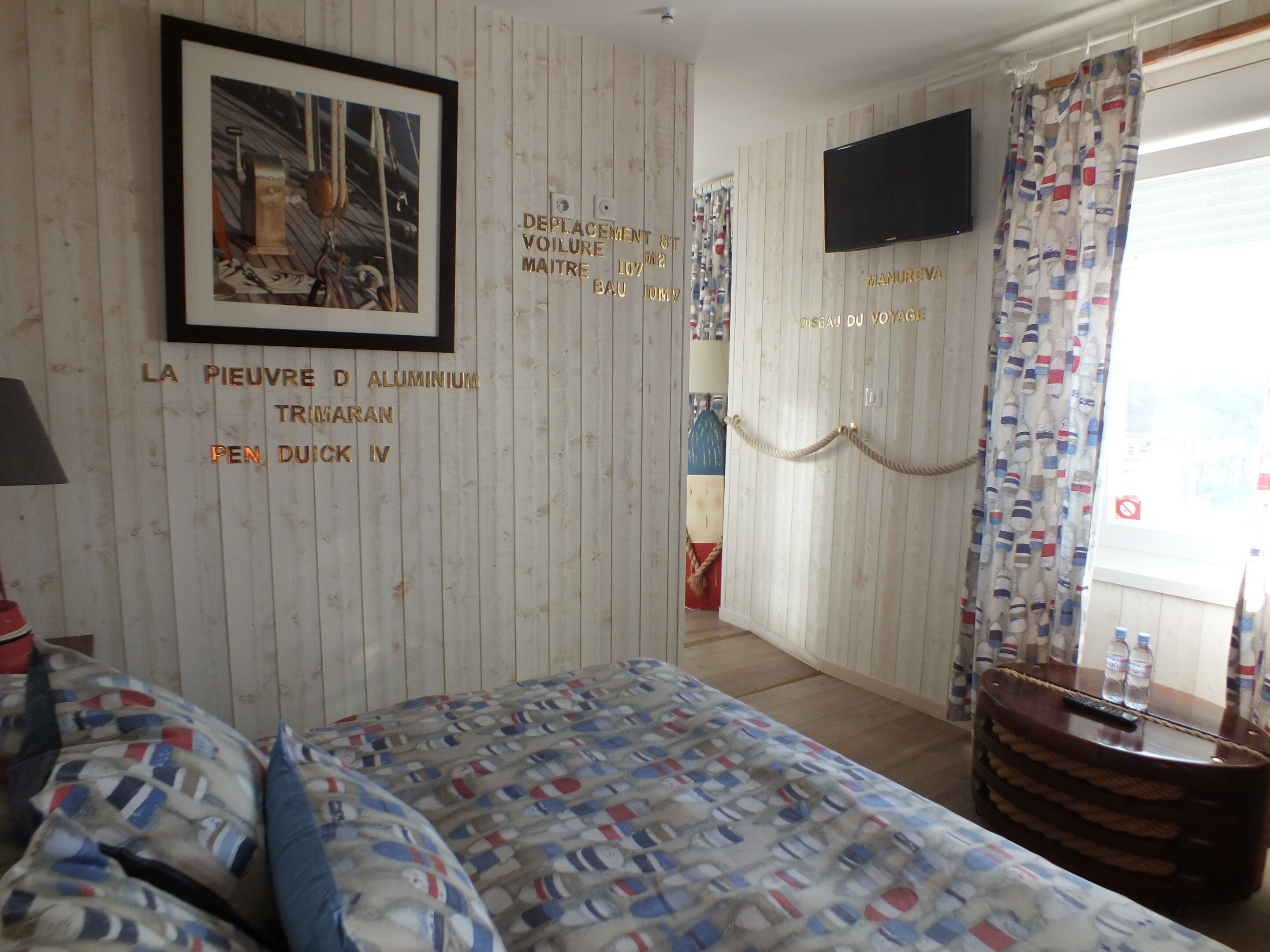 Une chambre d’hôtel avec un lit, une télévision, un rideau orné d’un motif représentant des bouteilles, et un mur avec du texte et un tableau encadré.
