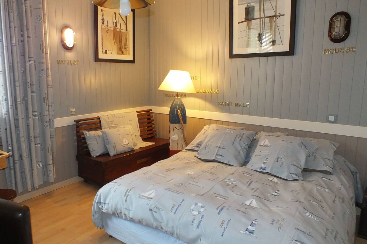A hotel room with a bed, a wooden chair, a desk with a chessboard, and a lamp.