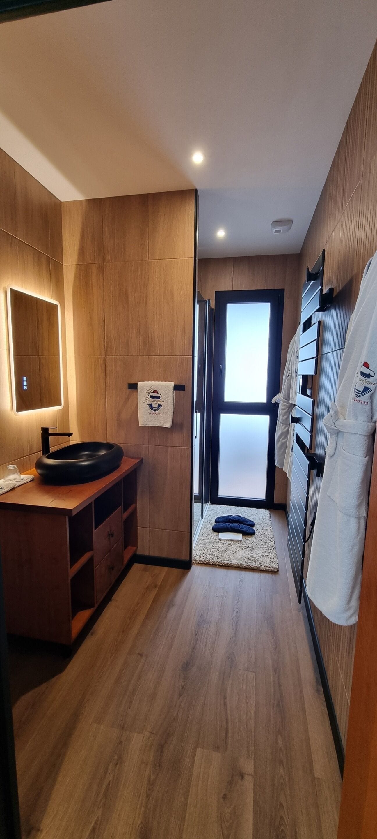 Une salle de bain moderne avec un meuble-lavabo en bois, un lavabo rond, un porte-serviettes et une vue sur l’extérieur grâce à une porte en verre.