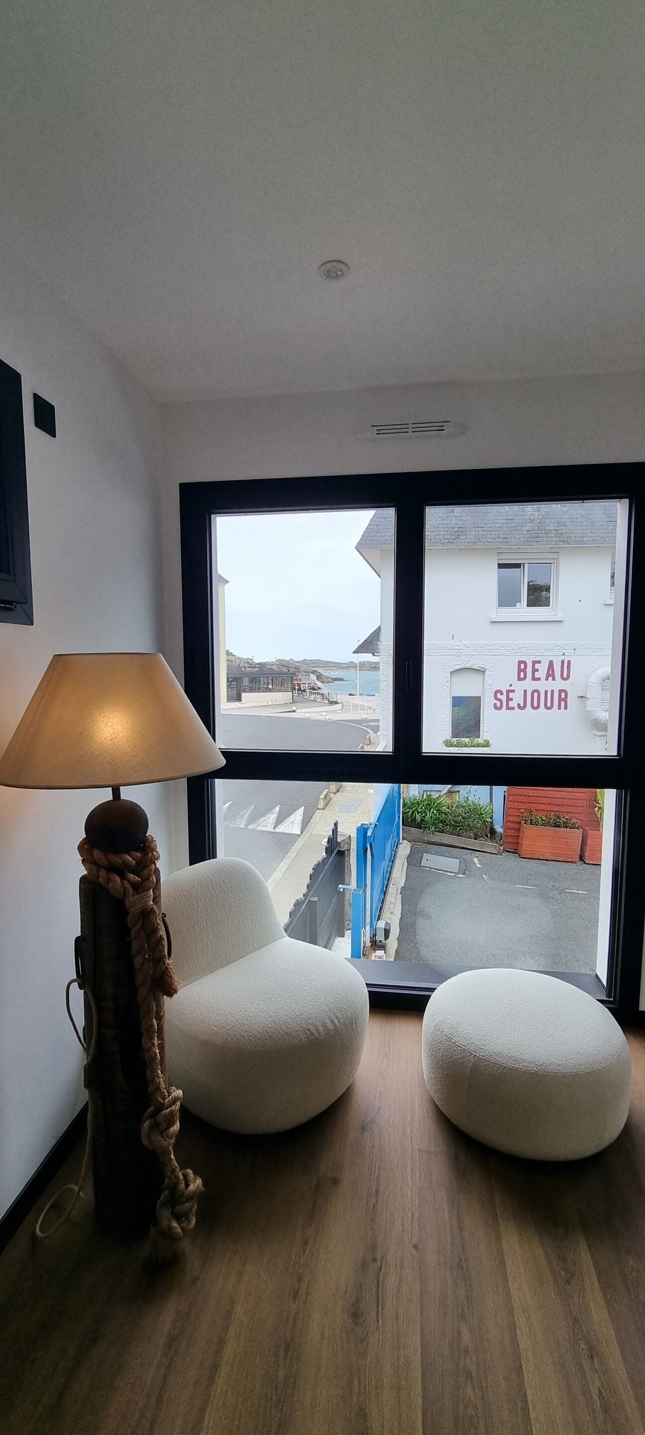A room with a large window, a lamp, two white chairs, and a wooden floor.
