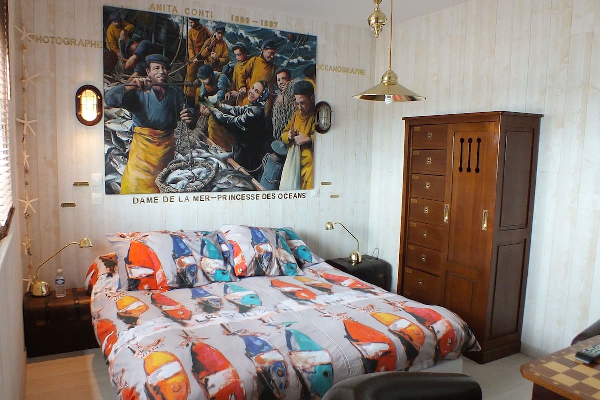 A bedroom with a bed featuring a colorful fish pattern, a wooden wardrobe, a large ocean-themed painting, and a chandelier.