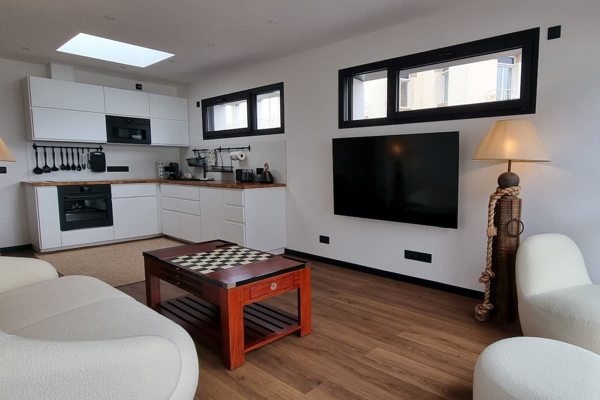 A modern living room with a wooden coffee table, white sofas, a kitchen area with white cabinets, a flat-screen TV, and two lamps with rope bases.