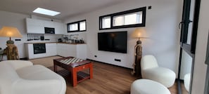 A modern living room with a wooden coffee table, white sofas, a kitchen area with white cabinets, a flat-screen TV, and two lamps with rope bases. - Le Beauséjour (Tregastel)