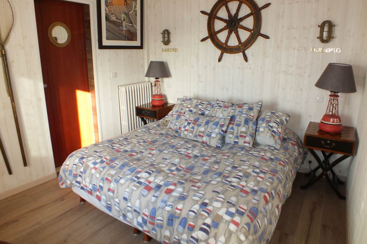 A bedroom with a bed, two bedside tables, a wooden door, a radiator, and a wall-mounted ship's wheel.