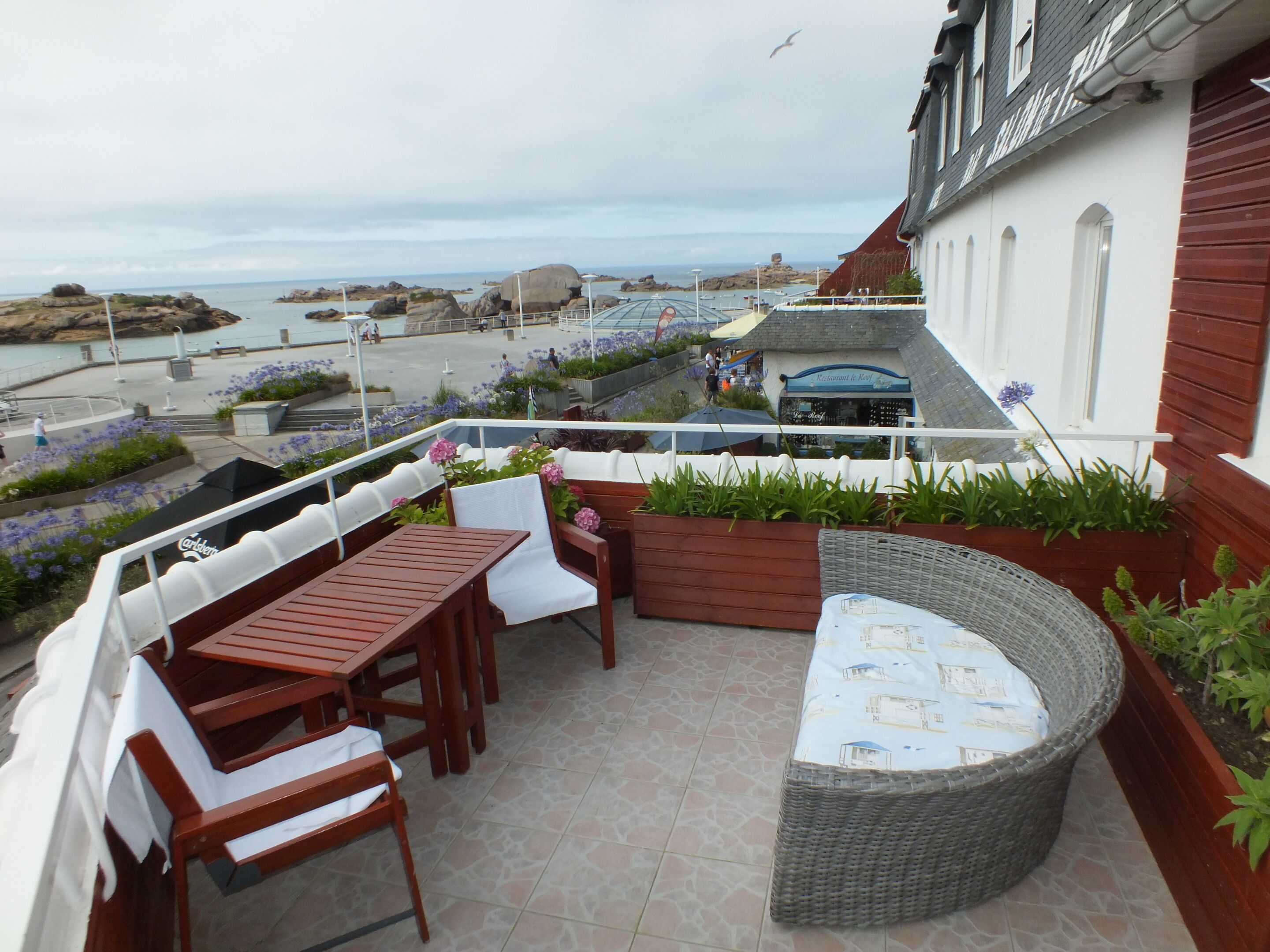 Une terrasse dotée de mobilier en bois, d’un canapé en osier et offrant une vue sur la mer et le paysage rocheux.