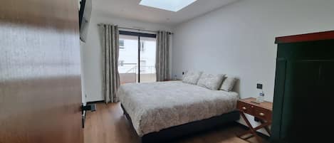 A bedroom with a bed, a nightstand, a desk, a chair, a window with curtains, and a skylight.