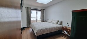 Un dormitorio con cama, mesita de noche, escritorio, silla, ventana con cortinas y tragaluz.