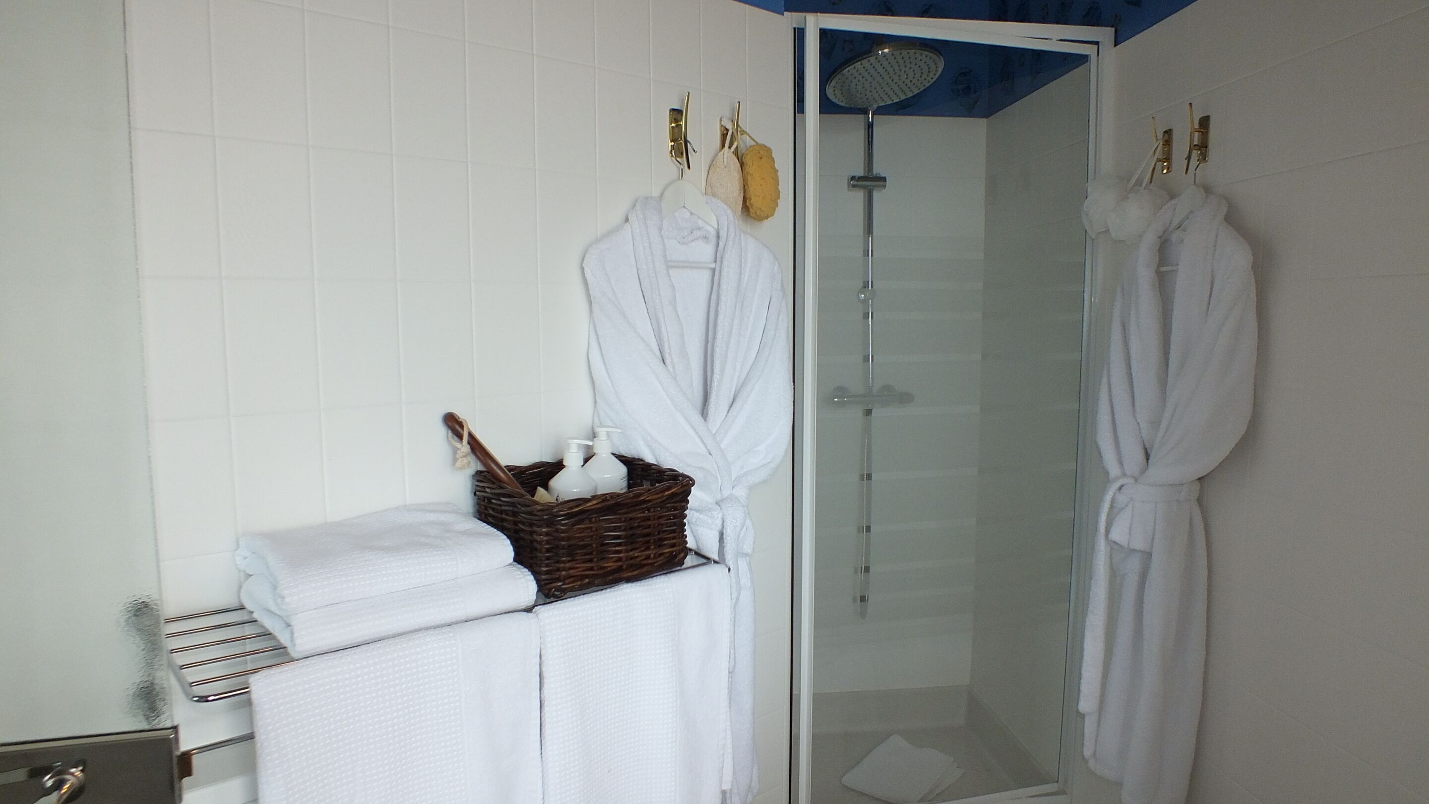 A bathroom with white tiled walls, a glass shower enclosure, and two white bathrobes hanging on hooks.