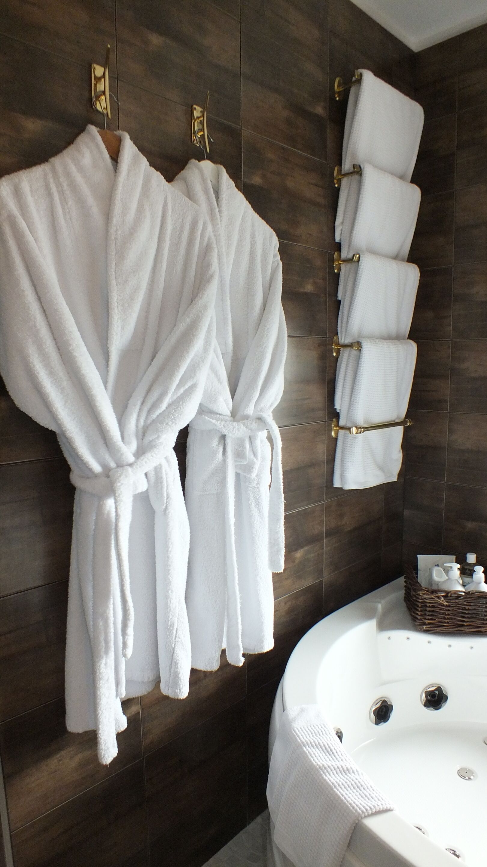 Un baño con dos albornoces blancos colgados en ganchos, una bañera blanca y una cesta de mimbre con artículos de tocador.