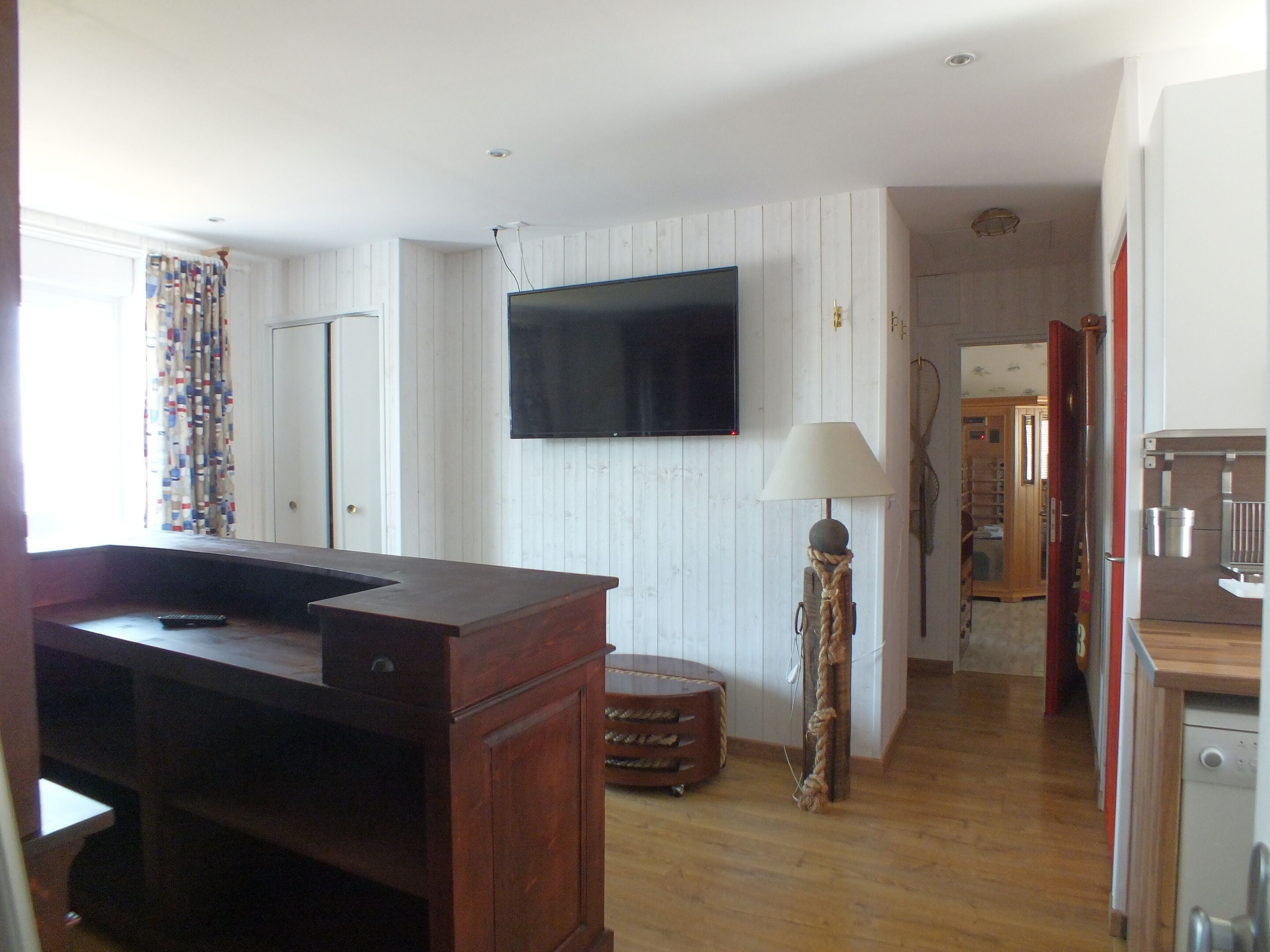 A room with a wooden floor, a television mounted on the wall, a lamp, and a kitchen area with a refrigerator.