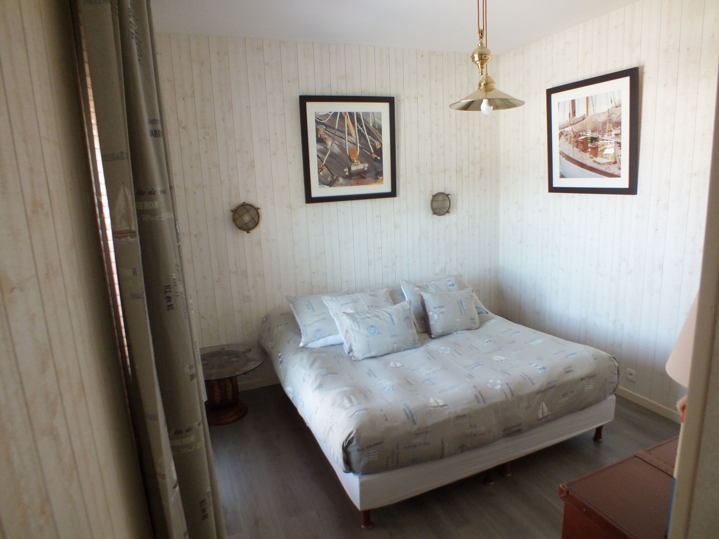 Ein Schlafzimmer mit einem Bett, zwei gerahmten Bildern an der Wand, einer Pendelleuchte und einem kleinen runden Tisch.