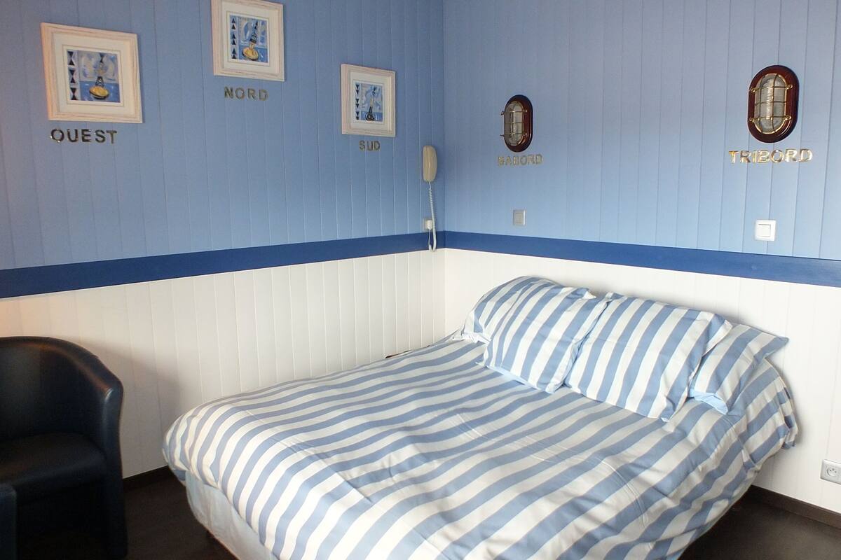 A single bed with striped bedding, a chair, and a wall with labeled sections 'NORD,' 'SUD,' 'BABOIRE,' and 'TRIBORD.'