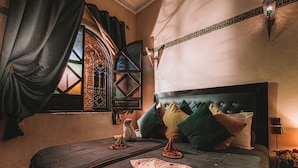 Un lit avec une tête de lit sombre, une fenêtre ornée de ferronnerie décorative et une lampe fixée au mur.