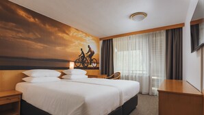 A hotel room with a large bed, a desk, a chair, and a wall mural of a sunset with cyclists.