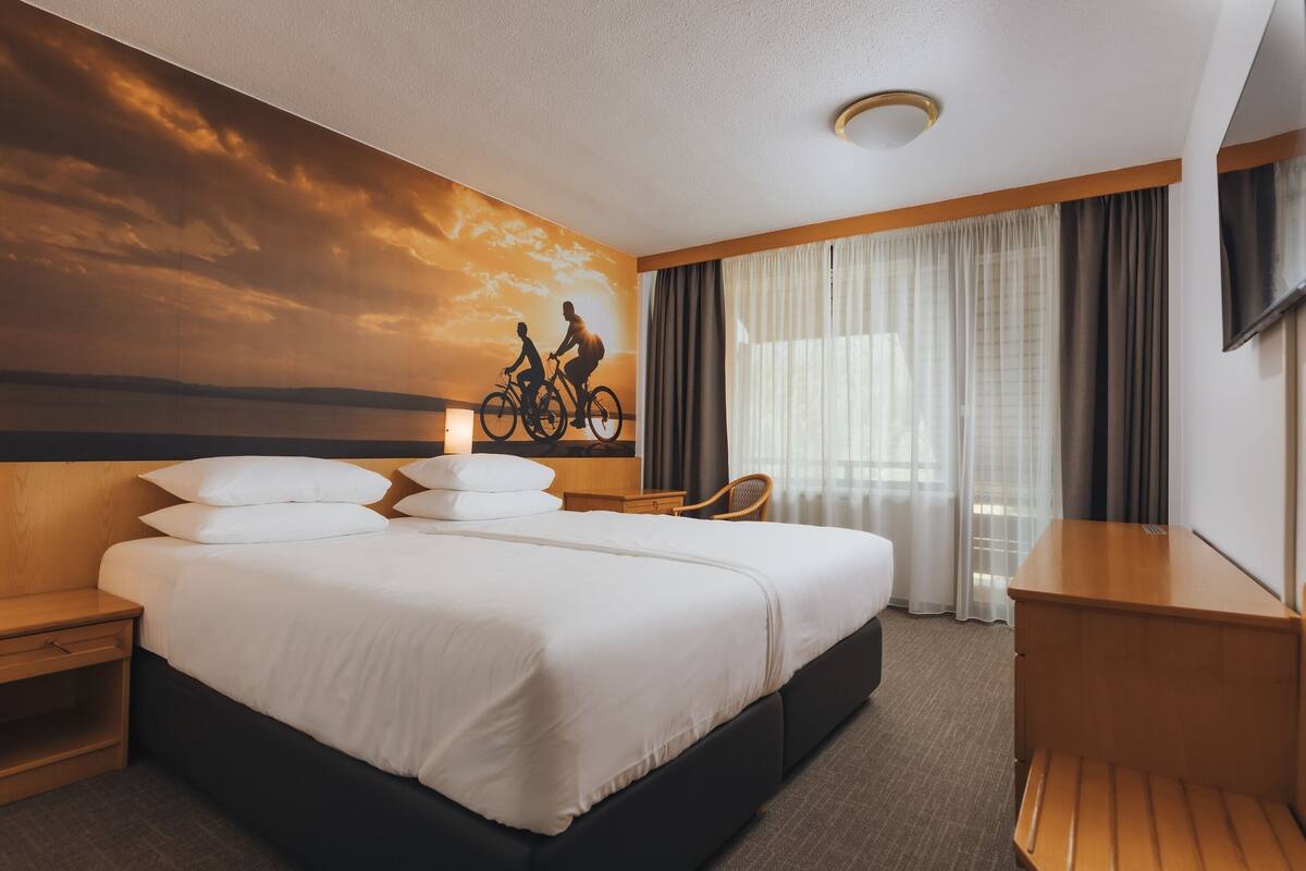 Habitación de hotel con una cama grande, un escritorio, una silla y un mural en la pared de un atardecer con ciclistas.