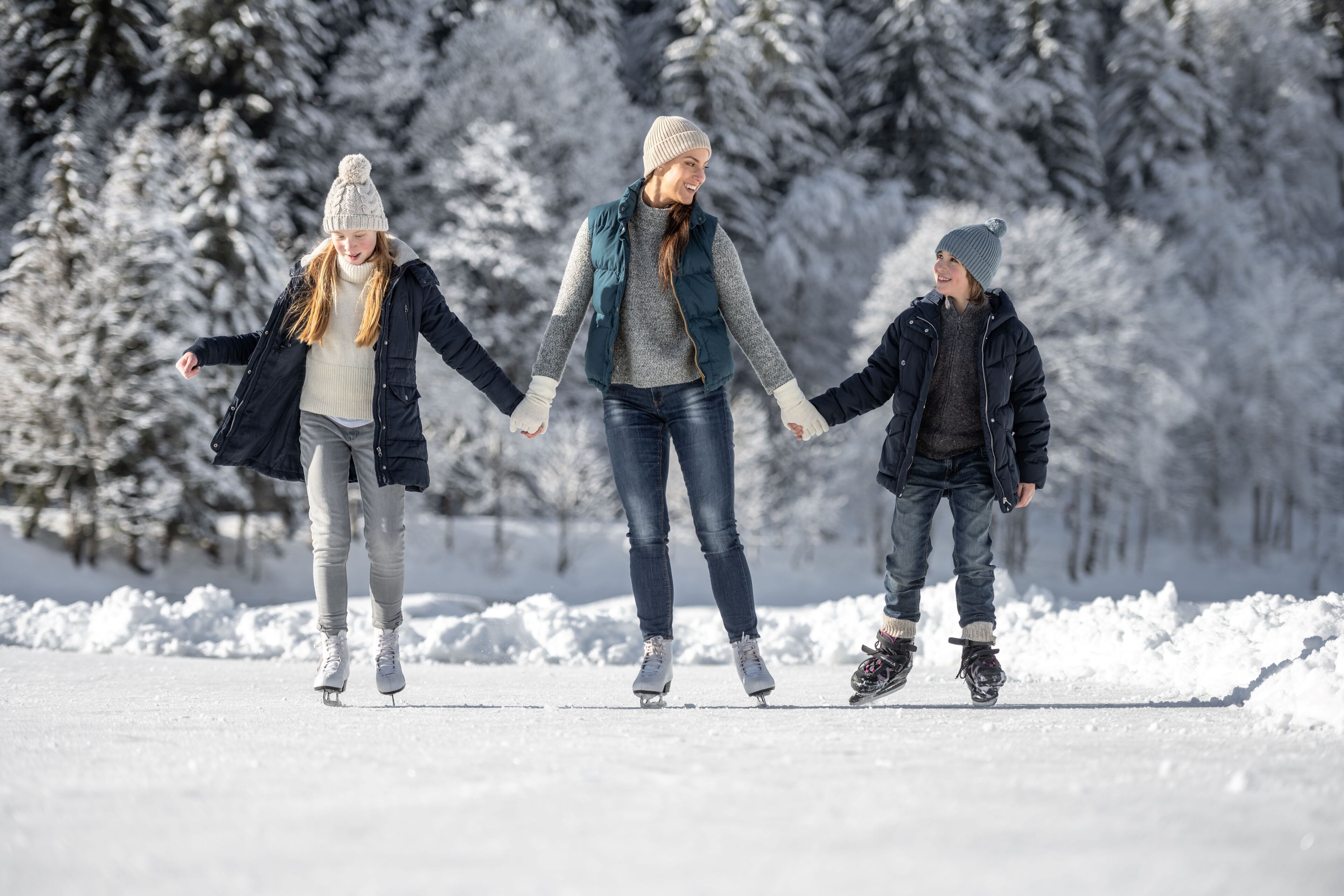 Three people ice skating on a frozen lake surrounded by snow-covered trees.