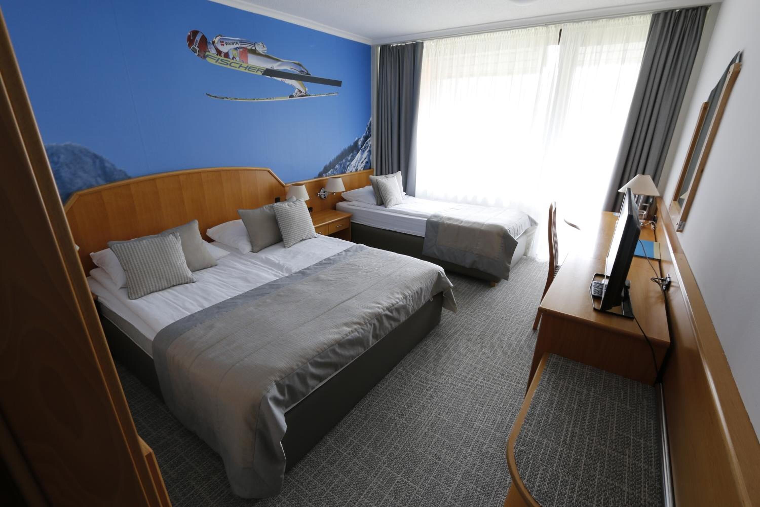 Habitación de hotel con una cama grande, una cama más pequeña, un escritorio con computadora, un espejo y un mural en la pared con temática de esquí.
