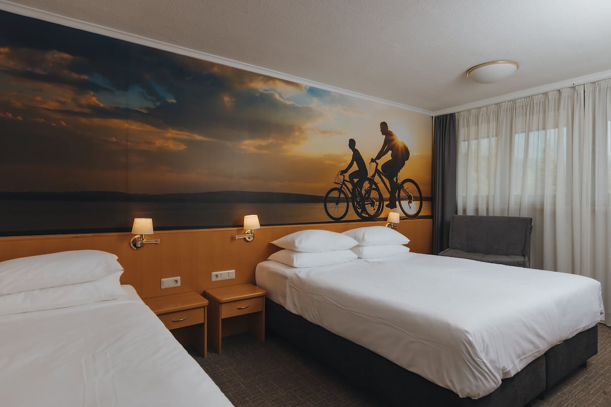 Habitación de hotel con dos camas, una mesita de noche, una lámpara, un mural en la pared y una ventana con cortinas.