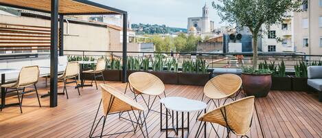 A wooden deck with wicker chairs and a table, overlooking a cityscape with buildings and a tree.