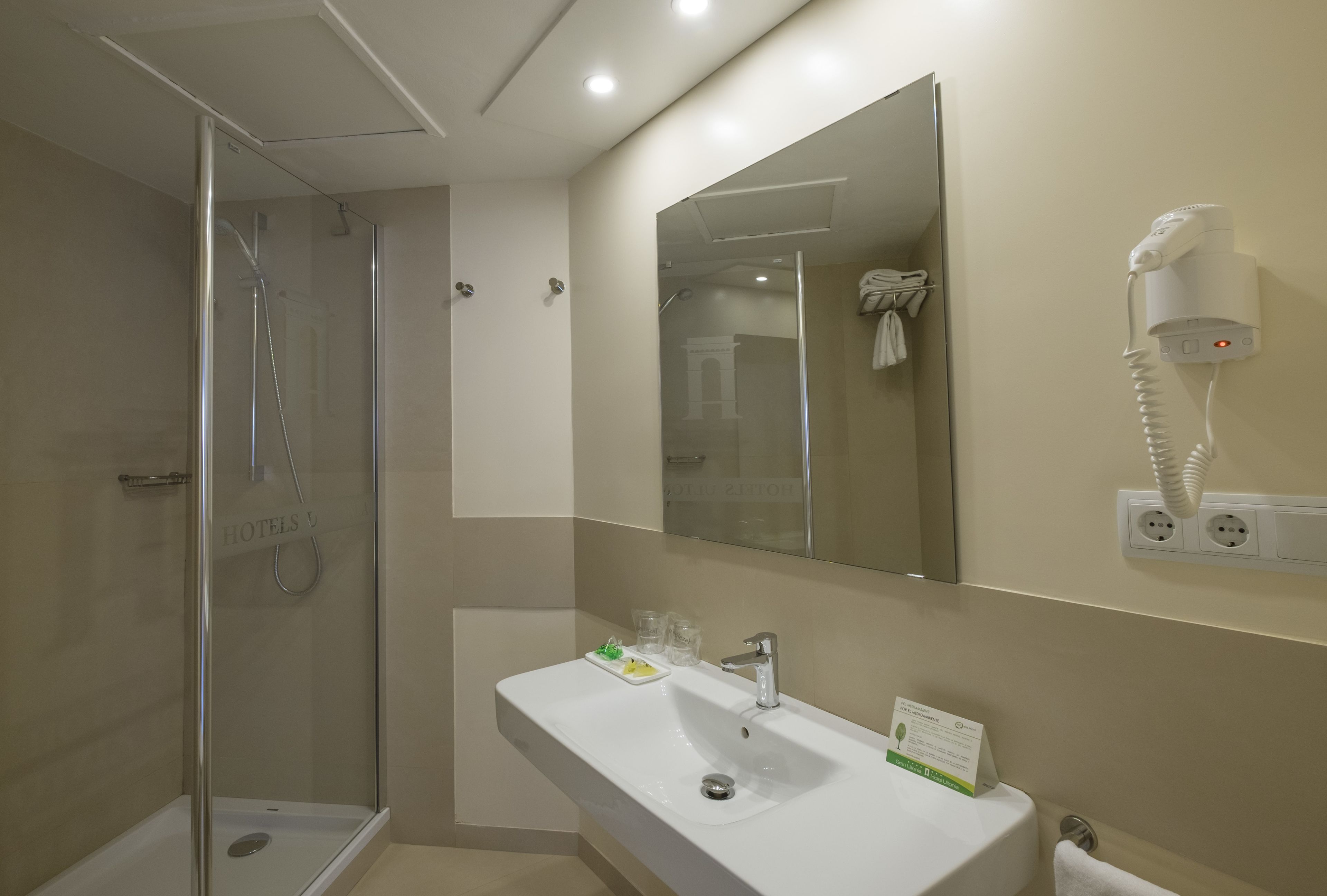 Un baño de hotel con ducha de vidrio, un lavamanos con espejo y un secador de pelo montado en la pared.