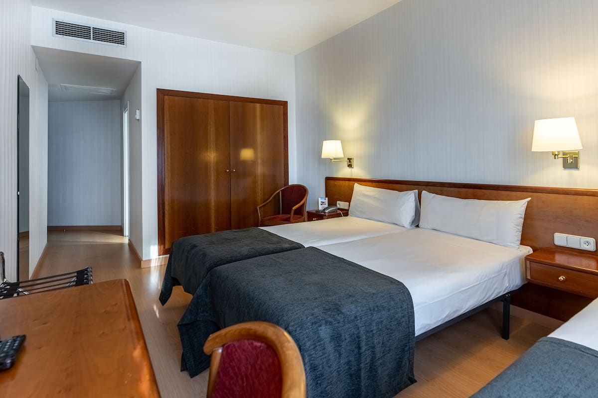 A hotel room with two beds, a desk with a chair, a wardrobe, and a bedside table with a lamp.
