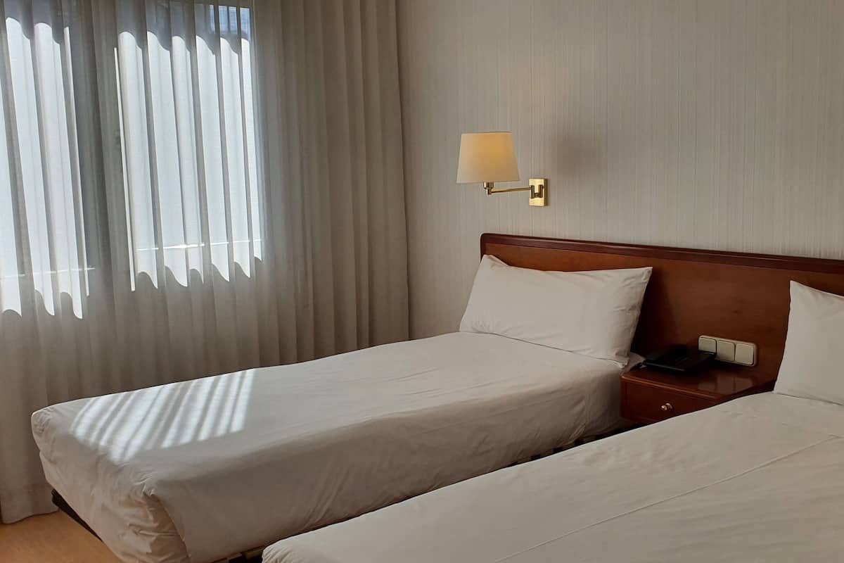 A hotel room with two single beds, a wooden headboard, a nightstand with a phone, and two wall-mounted lamps.