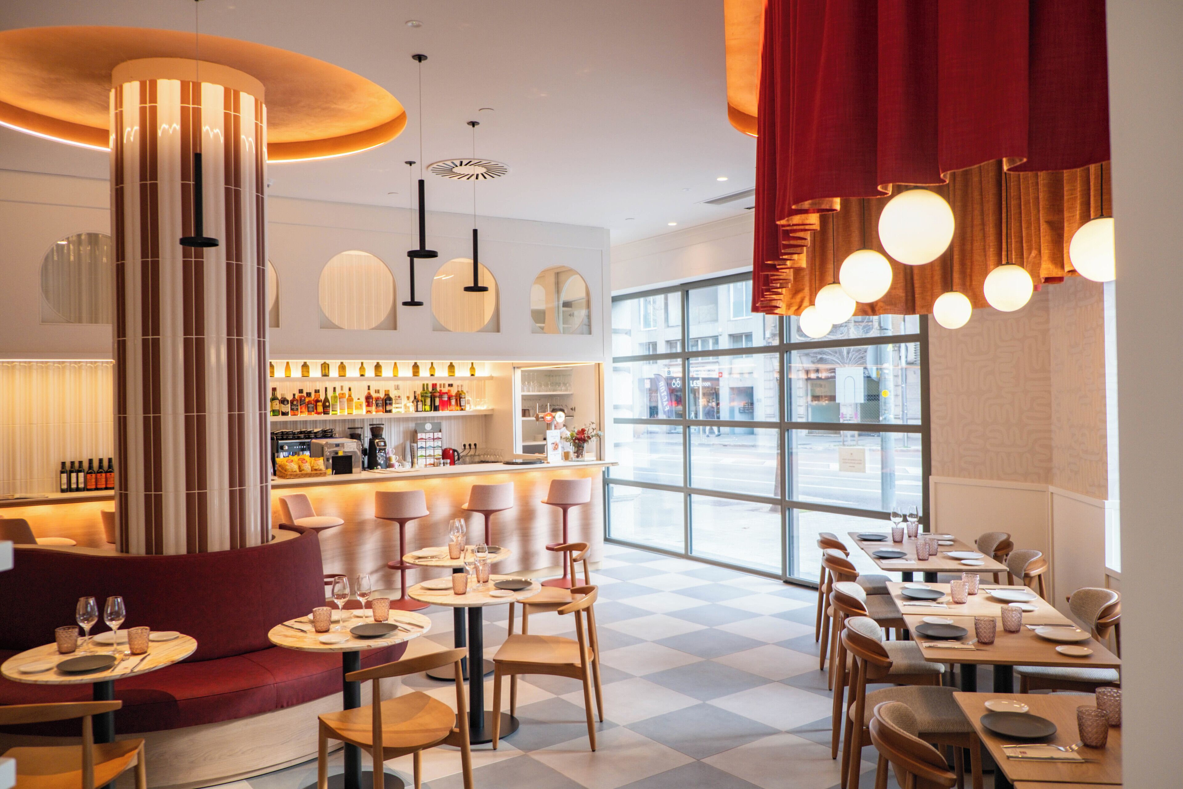 A modern restaurant interior with a bar area, seating arrangements, and large windows providing a view of the street.