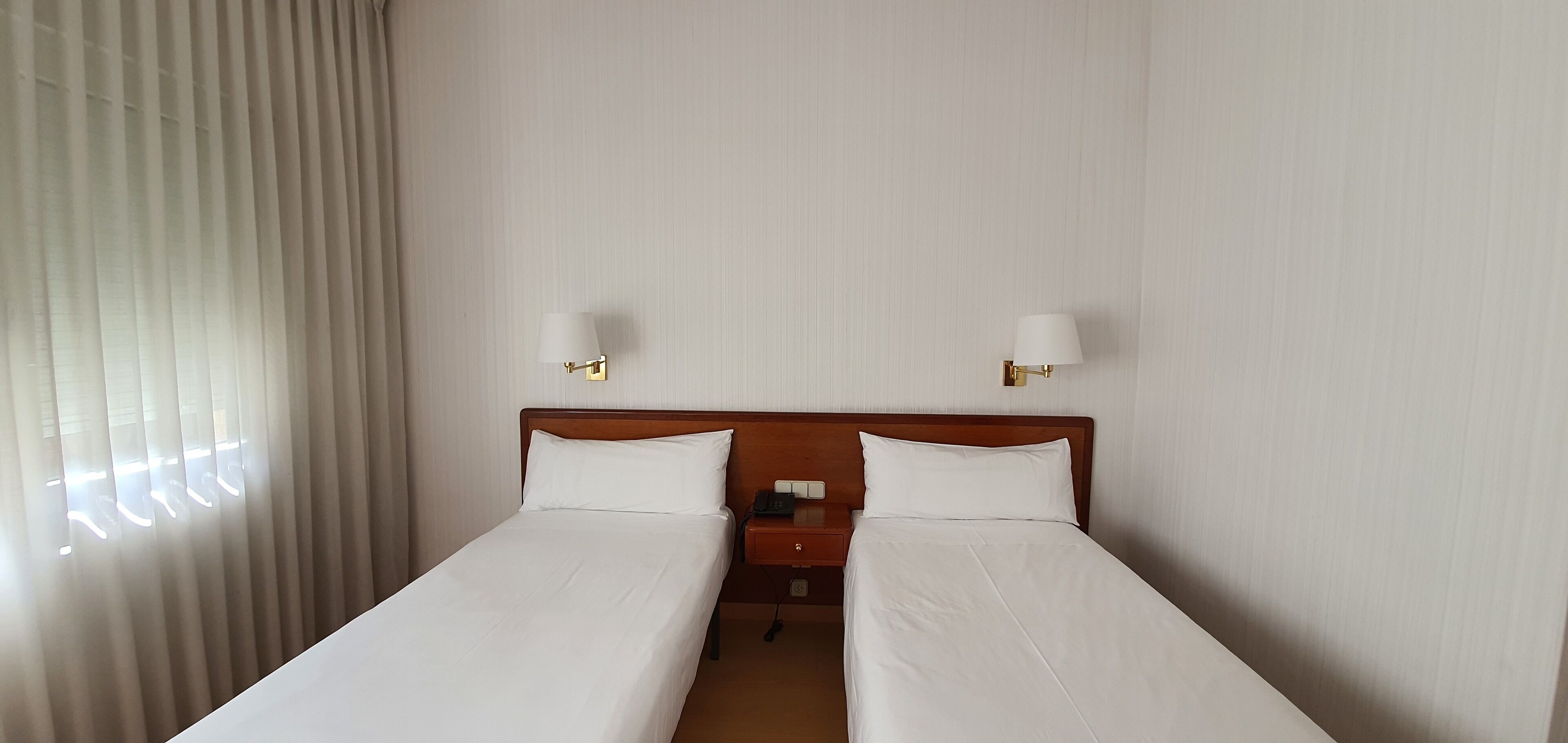 A hotel room with two single beds, a wooden headboard, a nightstand with a phone, and two wall-mounted lamps.