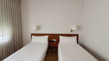 A hotel room with two single beds, a wooden headboard, a nightstand with a phone, and two wall-mounted lamps.
