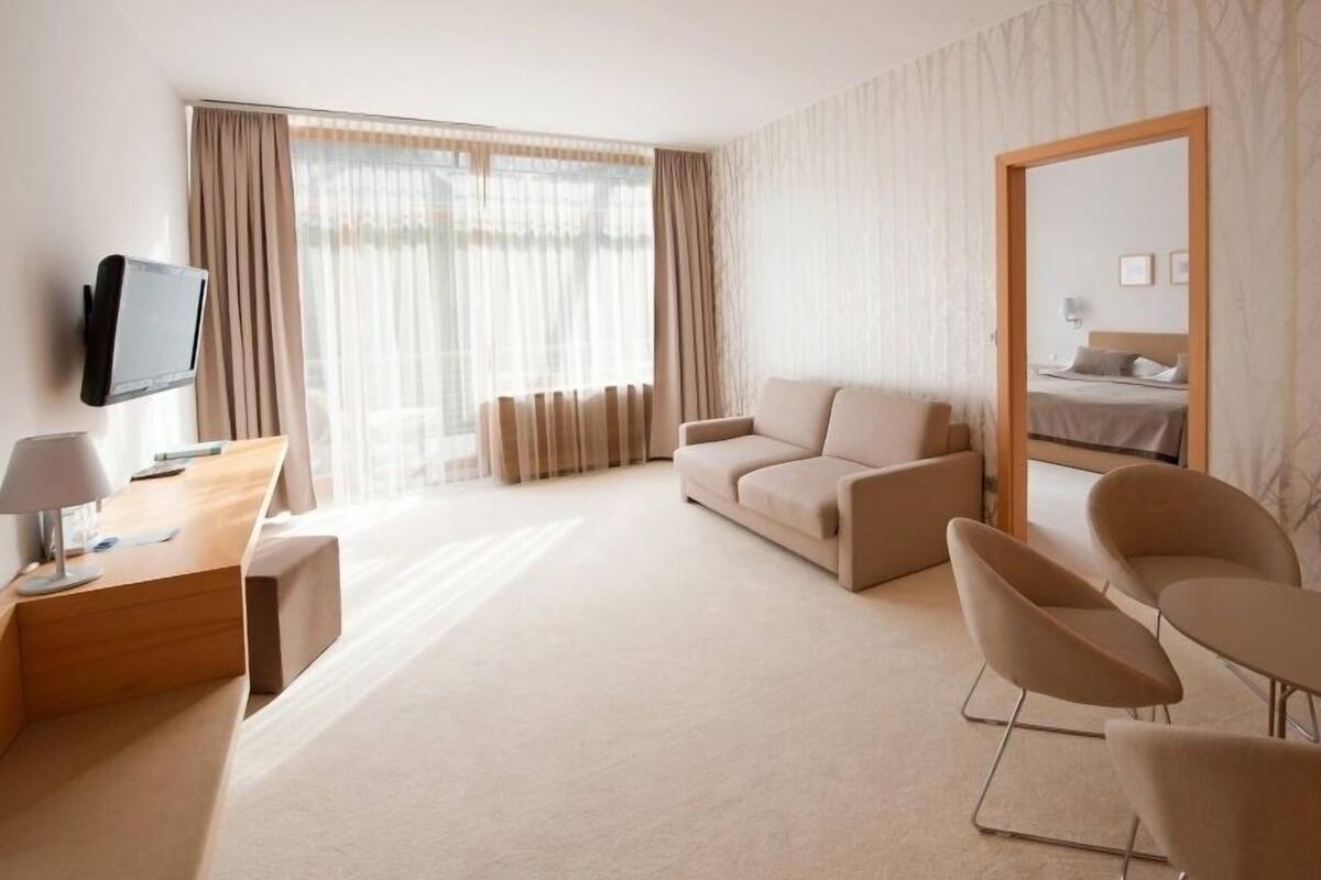 A modern hotel room with a sofa, two armchairs, a desk with a lamp, a television, and a large window with curtains.