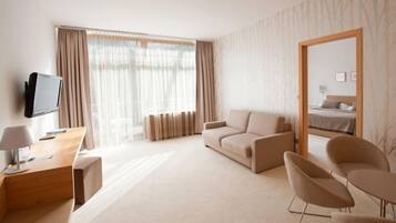 Una habitación de hotel moderna con un sofá, dos sillones, un escritorio con lámpara, televisión y un ventanal con cortinas.