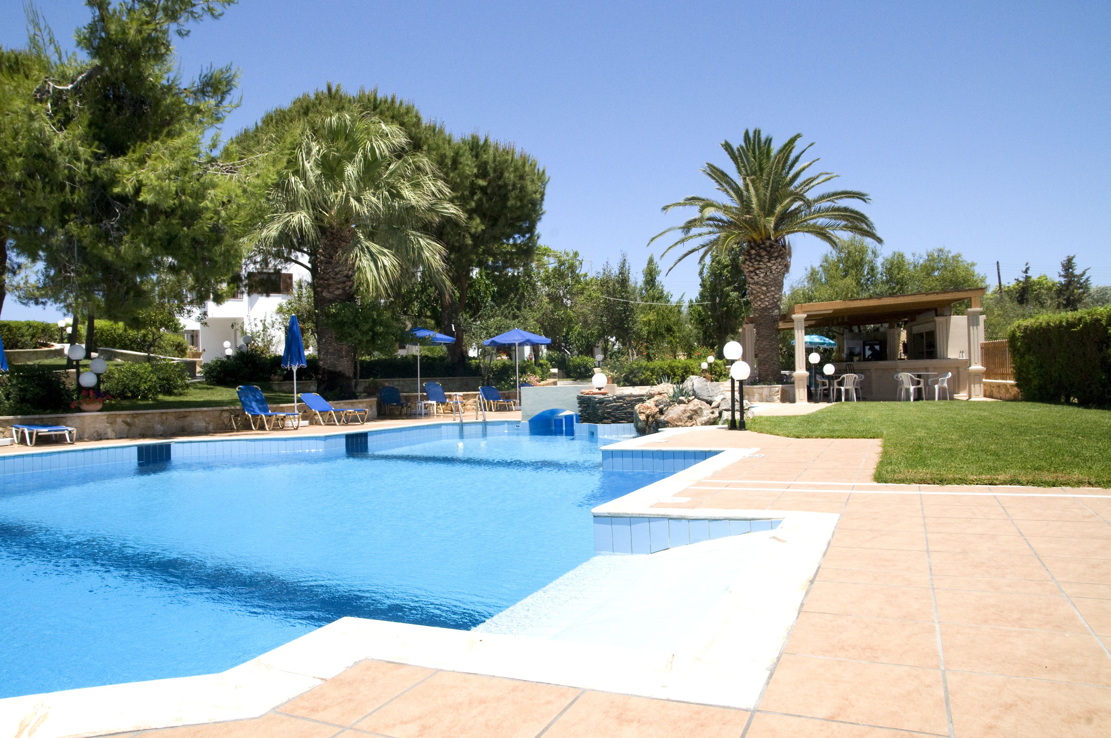 Una piscina con azulejos azules, rodeada de sillas de descanso y sombrillas, con palmeras y un edificio al fondo.