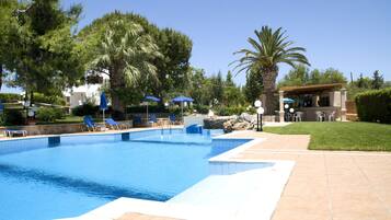 Una piscina con azulejos azules, rodeada de sillas de descanso y sombrillas, con palmeras y un edificio al fondo.