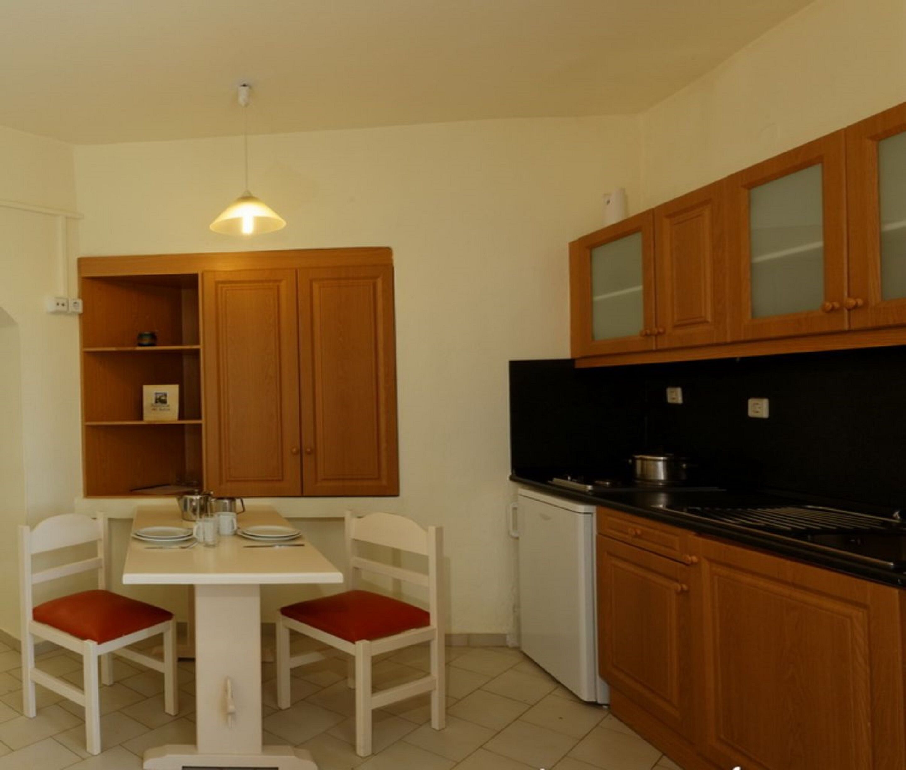 A compact kitchen with wooden cabinets, a white dining table with red chairs, and a small refrigerator.