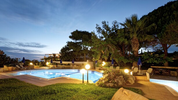 Una piscina con sillas de descanso y sombrillas, rodeada de árboles e iluminada por luces nocturnas.