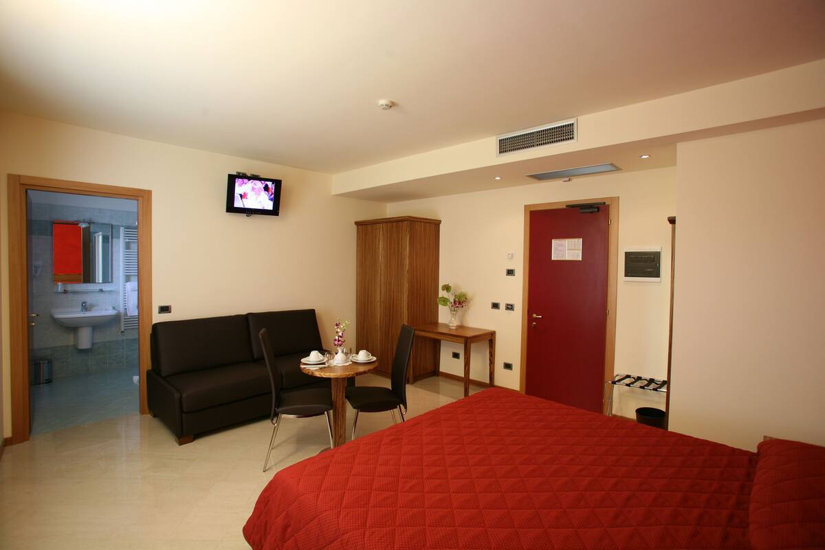 Ein Hotelzimmer mit einem Bett, einem Sofa, einem kleinen Tisch mit einer Vase, einem Fernseher an der Wand und einer Tür mit rotem Griff.