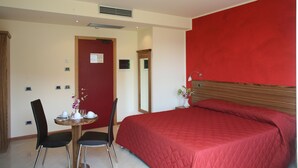 A hotel room with a red wall, a wooden headboard, a bed with a red quilt, a small round table with chairs, and a mirror.