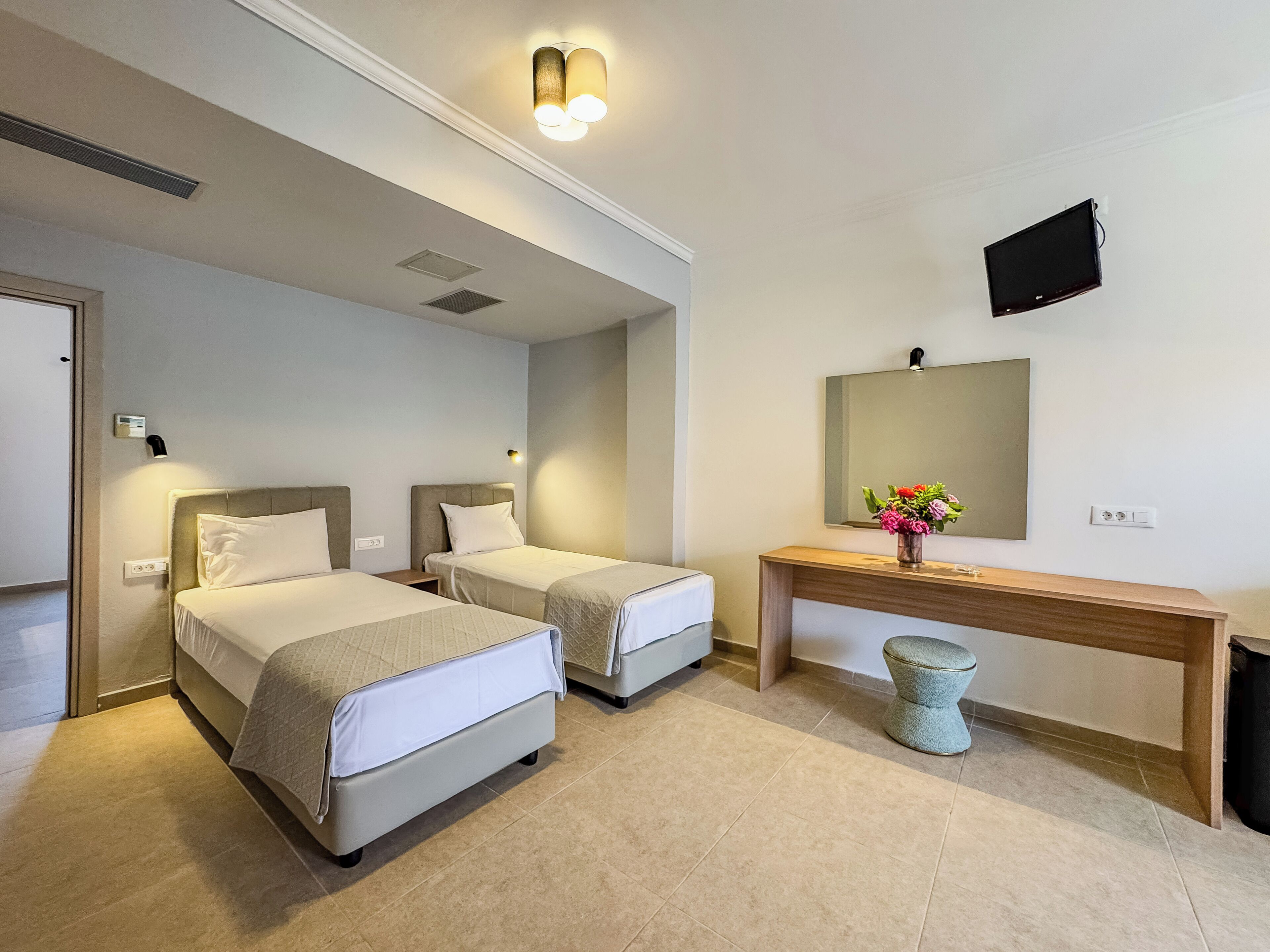 A hotel room with two beds, a small table with a vase of flowers, a television mounted on the wall, and a door.
