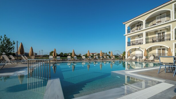 A pool area with lounge chairs and umbrellas, a two-story building with balconies, and clear blue skies. - Athina Apts (Zakynthos)