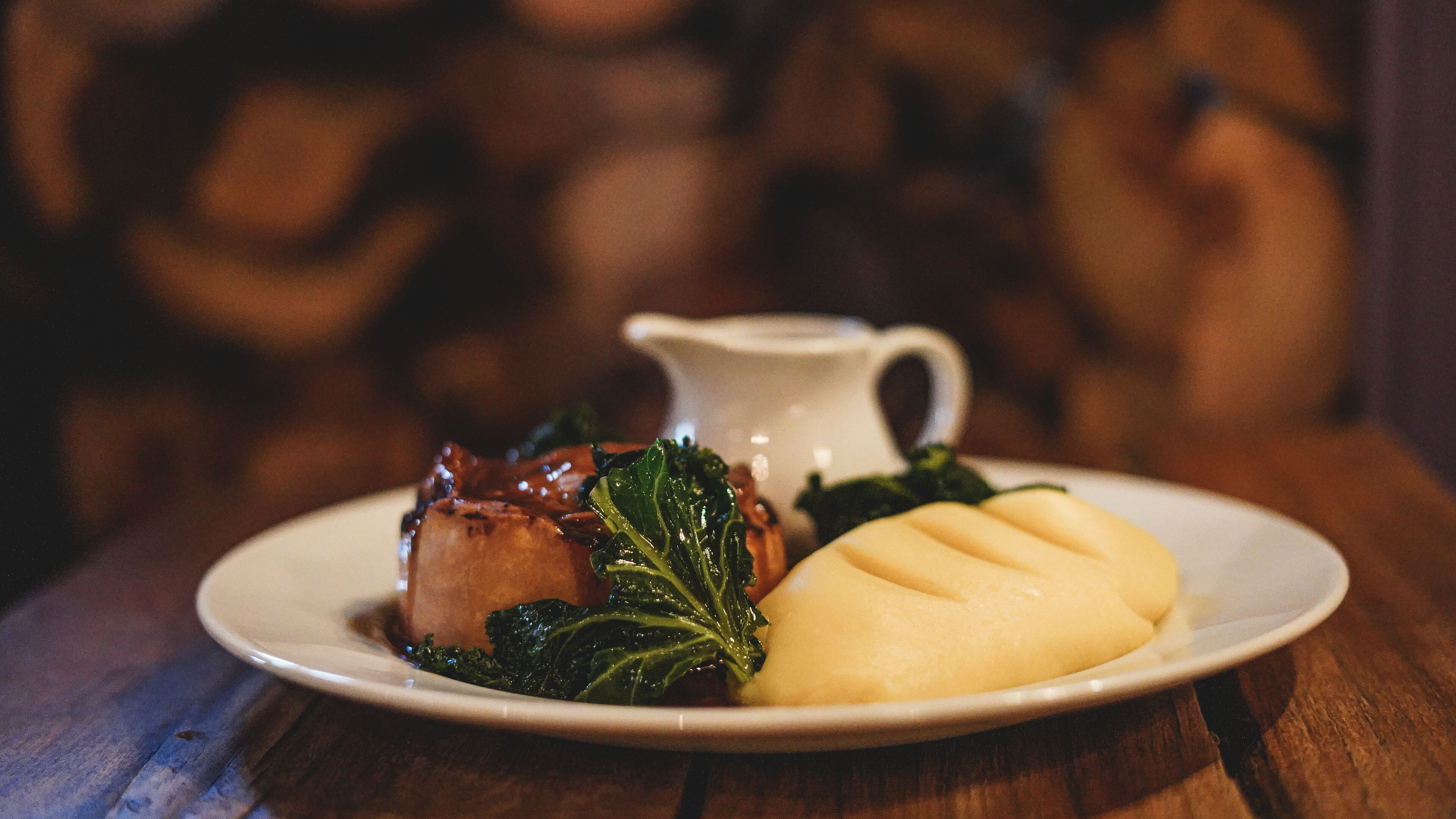 A plate with a piece of meat, mashed potatoes, and greens, accompanied by a small pitcher.